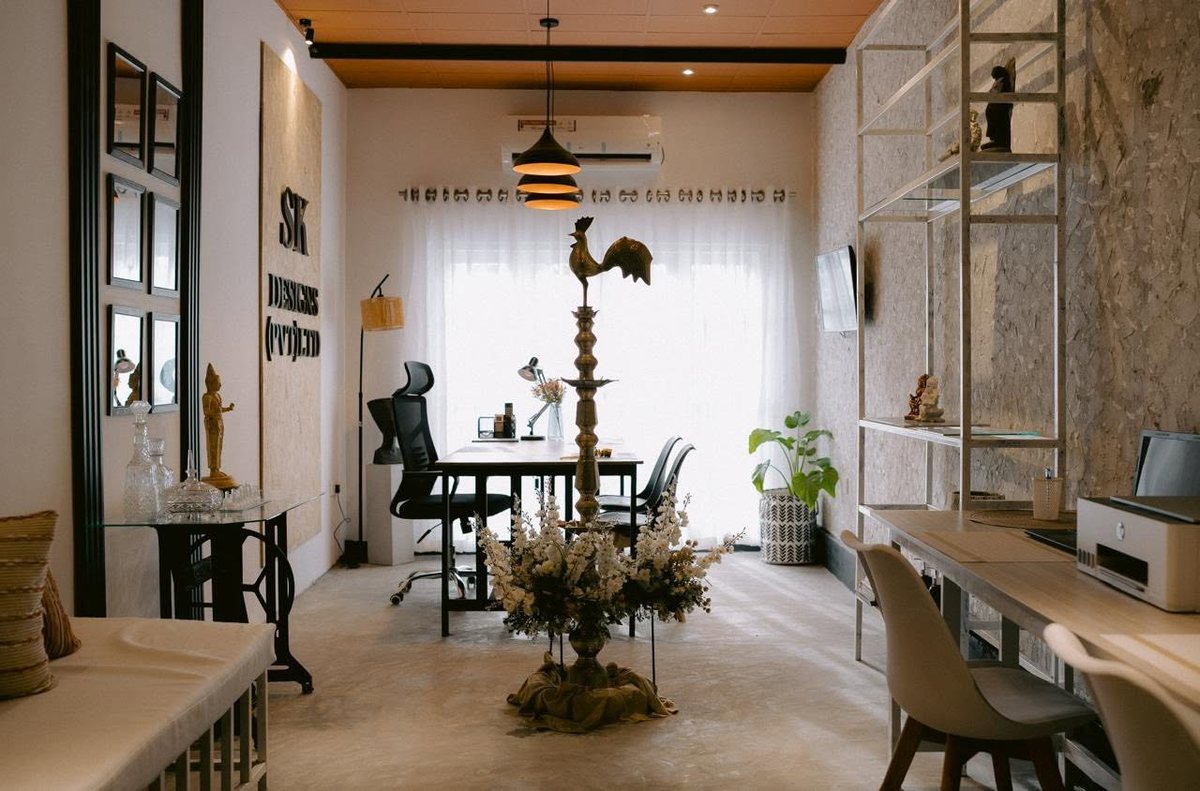 The main studio room, a brass oil lamp and flowers at the centre, desks against sheer curtained glazing, mirrors along the left wall and open steel shelving on the right