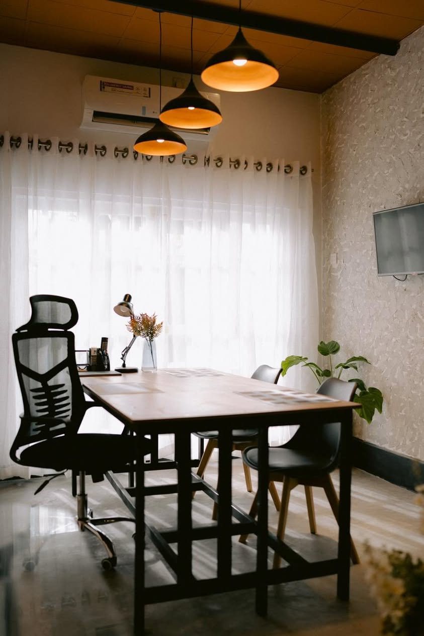 A meeting room with three black pendant lights over a timber table, sheer curtains across the window wall and a textured render finish