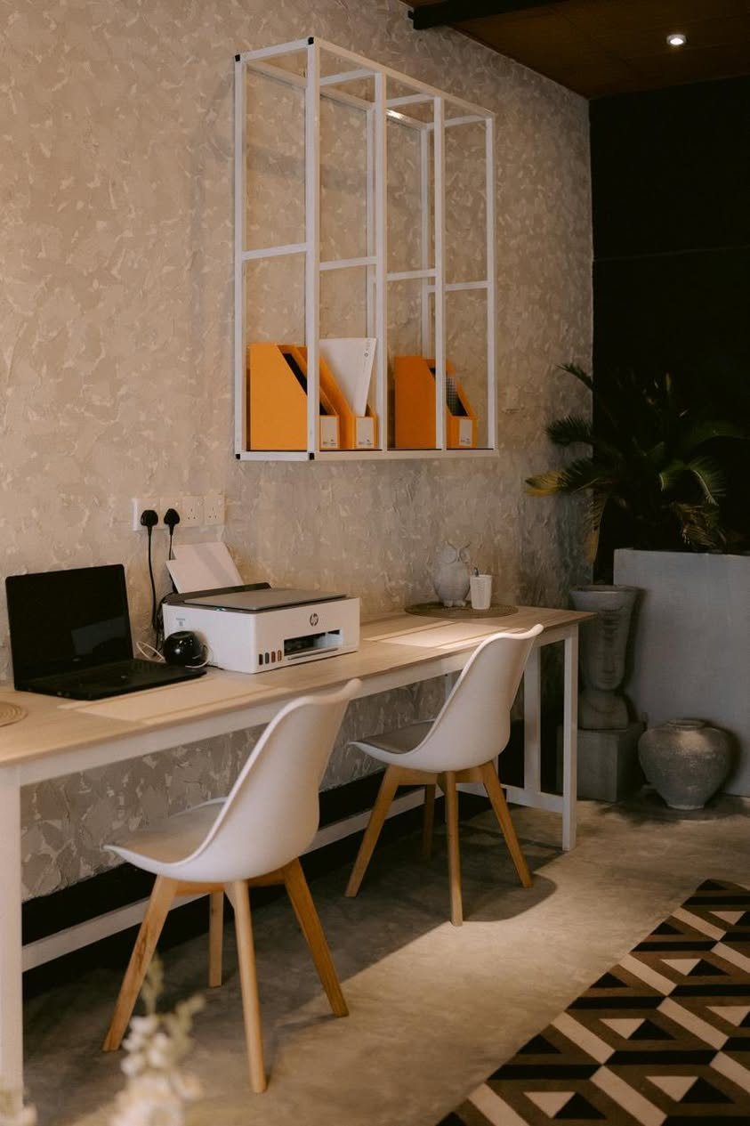 A long timber work desk with a printer and laptop, two white moulded chairs, a white steel wall rack and a geometric rug